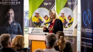 Woman with blonde hair, black jacket and red top stands at a podium at event talking to audience