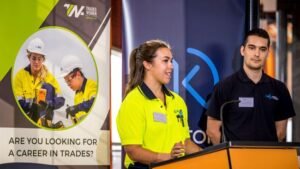 Apprentice in hi-vis standing next to man with navy polo top standing on podium. Apprentice is speaking to unseen crowd.
