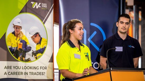 Apprentice in hi-vis standing next to man with navy polo top standing on podium. Apprentice is speaking to unseen crowd.